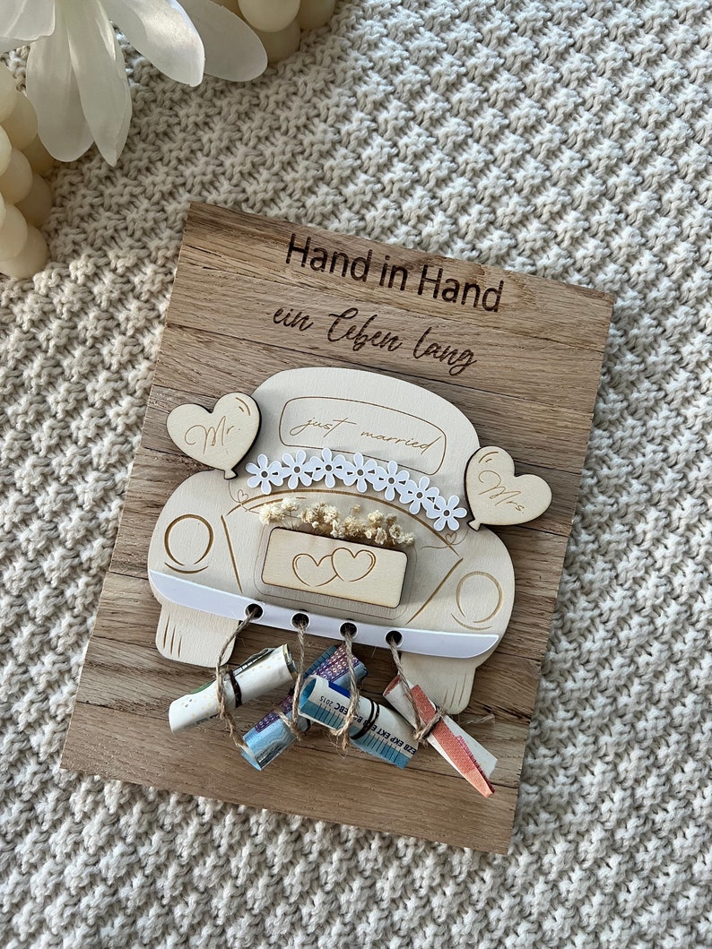 May include: A wooden wedding keepsake featuring a car design with the words "just married." The car is decorated with flowers and hearts, and the wooden plaque has the German text "Hand in Hand ein Leben lang." Several rolled-up bills are attached.