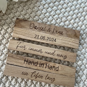 May include: Three wooden plaques with engraved text. The top plaque reads "Daniel & Lena" and "21.08.2024". The second plaque says "F&uuml;r immer und ewig". The bottom plaque says "Hand in Hand ein Leben lang".