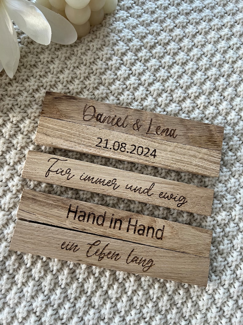 May include: Three wooden plaques with engraved text. The top plaque reads "Daniel & Lena" and "21.08.2024". The second plaque says "F&uuml;r immer und ewig". The bottom plaque says "Hand in Hand ein Leben lang".