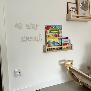 Könnte beinhalten: Eine weiße Wand mit Holzbuchstaben, die "es war einmal" buchstabieren, und ein Holzregal mit Büchern und einem Bilderrahmen. Das Regal ist an der Wand befestigt. Der Bilderrahmen hat einen braunen Rahmen und ein Bild eines Faultiers mit dem Wort "HEY" darunter.
