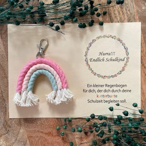 Op de afbeelding: Een roze, witte en groene regenboog sleutelhanger met witte kwastjes. De sleutelhanger heeft een zilveren clip. De tekst "Hurra!!! Endlich Schulkind" staat op een kaart achter de sleutelhanger. De tekst "Ein kleiner Regenbogen für dich, der dich durch deine kunterbunte Schulzeit begleiten soll." staat ook op de kaart.