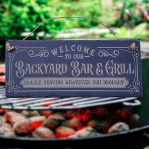 May include: A blue slate sign with white text that reads "Welcome to our Backyard Bar & Grill Gladly serving whatever you brought". The sign has a rope hanger and is hanging above a grill.