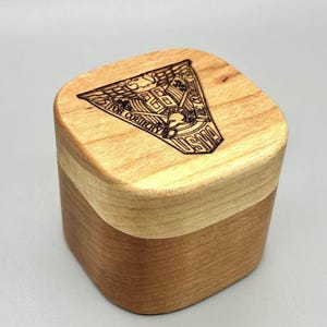 May include: A two-tone wooden box with a laser-engraved emblem on the lid. The emblem features text that reads "FOR COUNTRY" and "USMA". The box is square with rounded edges, and the wood has a natural finish.