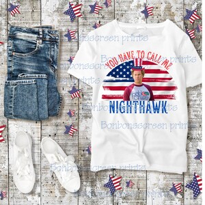 Puede incluir: Una camiseta blanca con el texto "YOU HAVE TO CALL ME NIGHTHAWK" sobre un diseño de bandera estadounidense y una foto de un hombre. La imagen también incluye vaqueros azules y zapatillas blancas.