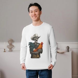 May include: White long-sleeve shirt featuring a skeleton wearing a cowboy hat and holding a rooster. The skeleton is dressed in a plaid shirt and jeans. The rooster is orange and brown. The shirt is worn by a person.