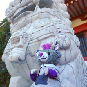 May include: A gray plush toy with purple accents is sitting in front of a large white marble lion statue. The toy has a purple and white hoodie and a purple tail.