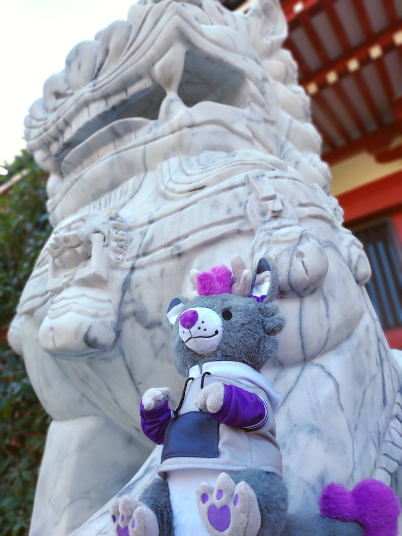 May include: A gray plush toy with purple accents is sitting in front of a large white marble lion statue. The toy has a purple and white hoodie and a purple tail.