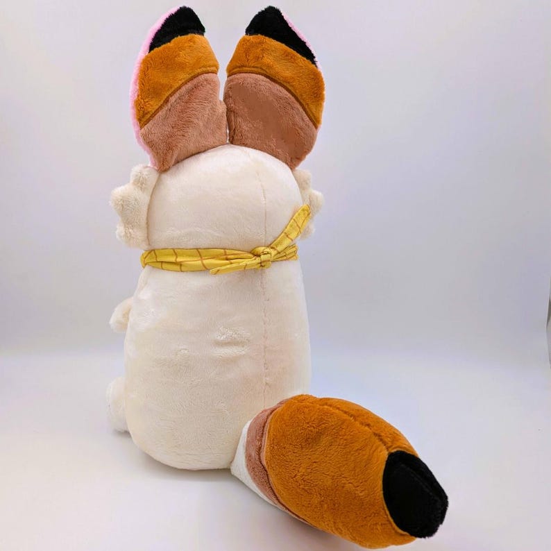 May include: A plush toy fox with large, multi-colored ears and a matching tail. The ears are pink, brown, and black. The fox's body is cream-colored, with a yellow scarf tied around its middle. The tail is brown, white, and black.