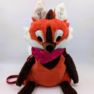 May include: A plush fox backpack in shades of orange, brown, and white, with a pink bandana featuring a floral pattern. The fox has large, expressive eyes and a red strap.