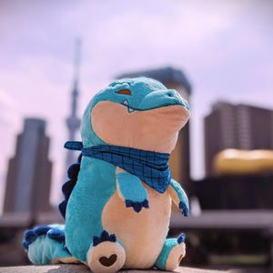 May include: A plush alligator toy in shades of blue and tan, with a blue bandana around its neck. The alligator has a heart-shaped detail on its foot and is sitting upright. The background is blurred, suggesting an outdoor setting.