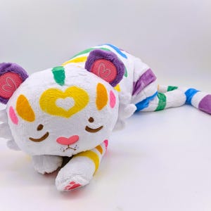 May include: A soft toy tiger in a relaxed pose, featuring a white body with rainbow stripes and colourful accents. The tiger has purple ears with pink heart details, a yellow heart on its forehead, and a pink nose. The toy is designed to be soft and cuddly.