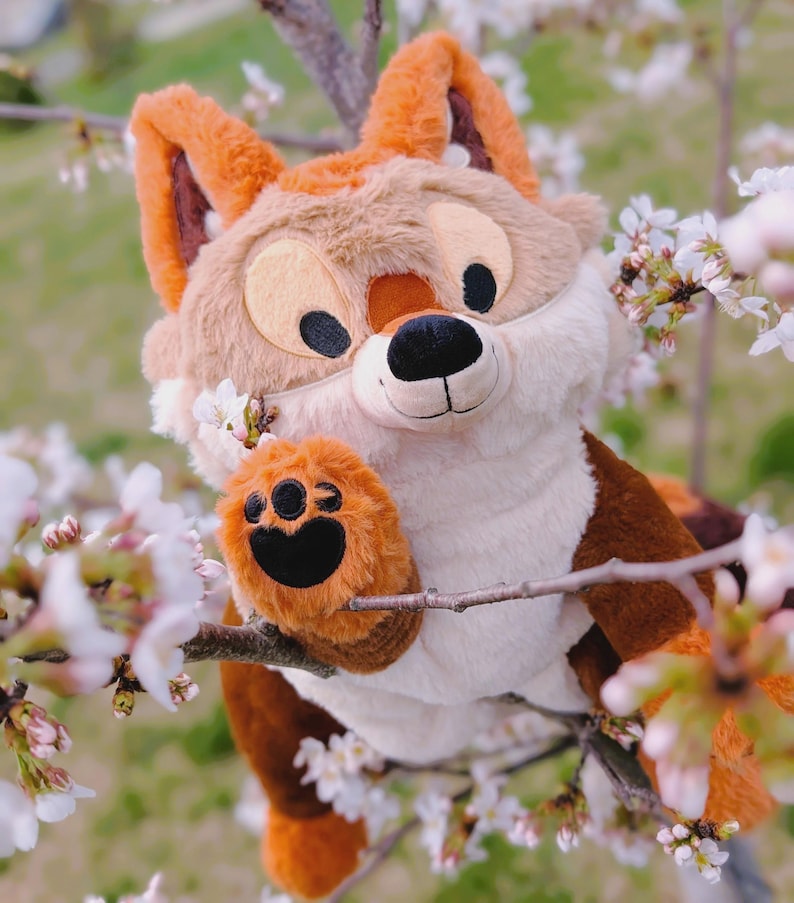 Puede incluir: Un peluche de zorro con pelaje marr&oacute;n, beige y blanco, ojos negros y nariz negra. El zorro est&aacute; posado en una rama con flores blancas. La pata del zorro est&aacute; extendida y el fondo est&aacute; borroso.