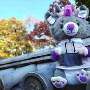 May include: A gray plush toy with purple accents, wearing a gray and white hoodie. The toy has a purple mohawk and antlers. The toy is sitting on a gray tiled surface.