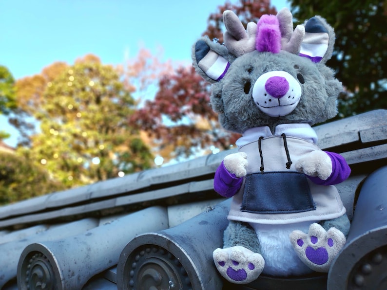 May include: A gray plush toy with purple accents, wearing a gray and white hoodie. The toy has a purple mohawk and antlers. The toy is sitting on a gray tiled surface.