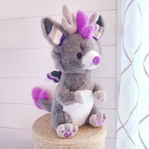 May include: A gray and purple plush toy dragon with a white belly and purple horns. The dragon is sitting on a woven basket.