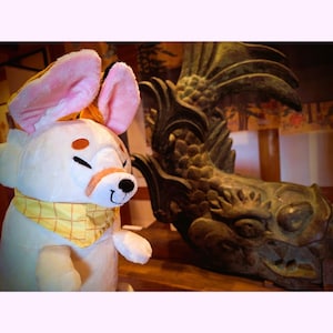May include: A plush white animal with large pink-lined ears, orange accents, and a yellow bandana. Beside it is a detailed bronze sculpture of a mythical creature, possibly a dragon or griffin, with intricate scales and feathers.
