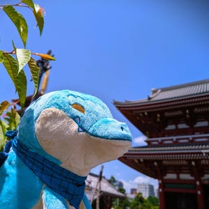 May include: A plush, blue and white crocodile toy with a blue bandana. The toy is set against a backdrop of a traditional Japanese building and a clear blue sky, with green leaves in the foreground.