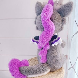 May include: A gray plush dragon with a purple tail and wings. The wings have a black, white, and purple design. The dragon is sitting on a woven basket.