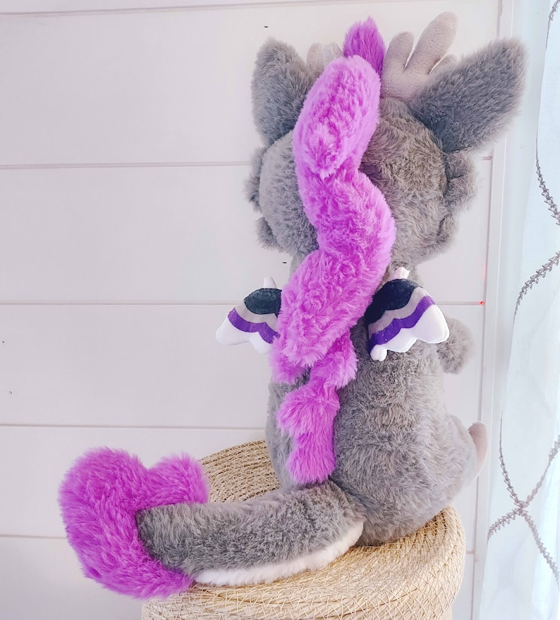 May include: A gray plush dragon with a purple tail and wings. The wings have a black, white, and purple design. The dragon is sitting on a woven basket.