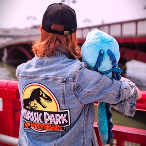 May include: A denim jacket with a Jurassic Park 30th Anniversary patch on the back. The jacket is worn with a black cap and a blue plush toy. The patch features a dinosaur silhouette and the film's logo.