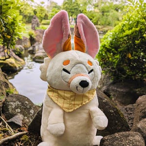 May include: A plush toy in the shape of a white animal with large pink and orange ears. It has orange eyes, a tan nose, and a yellow bandana. The toy is sitting on a rock near a body of water.
