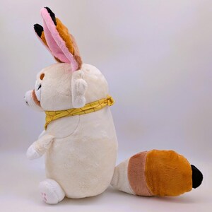 May include: A plush toy of a Fennec fox in a sitting position. The toy is primarily cream-colored with large, upright ears featuring pink and brown accents. It has a yellow bandana around its neck and a tail with brown, cream, and orange sections. The fox has small, white paws.