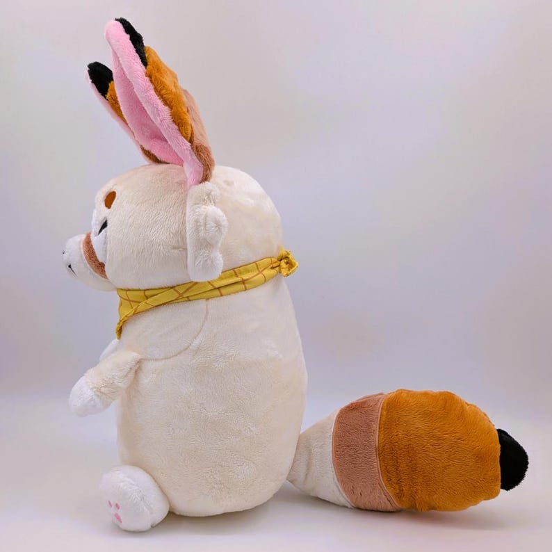 May include: A plush toy of a Fennec fox in a sitting position. The toy is primarily cream-colored with large, upright ears featuring pink and brown accents. It has a yellow bandana around its neck and a tail with brown, cream, and orange sections. The fox has small, white paws.