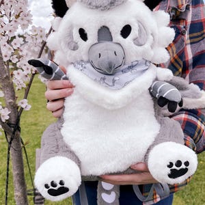May include: A white and gray plush backpack shaped like a mythical creature with a long tail, black eyes, and a gray beak. The backpack has a white bandana around its neck and a gray and white striped pattern on its back legs.