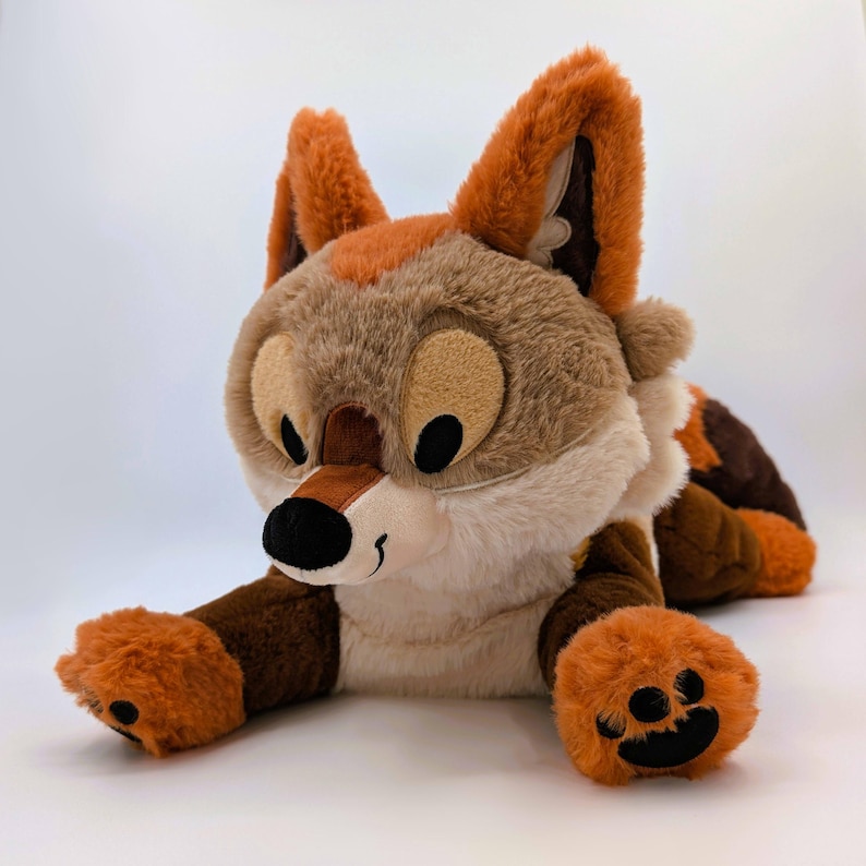 Puede incluir: Un peluche de zorro en tonos marr&oacute;n, naranja y crema, tumbado. El zorro tiene ojos grandes y redondos, una nariz negra y patas naranjas. Las orejas son erguidas y naranjas. El juguete es suave y tierno.