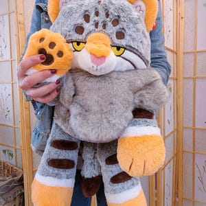 May include: A plush toy shaped like a wild cat with grey fur, dark brown spots on the head, and dark brown stripes on the limbs. The cat has yellow eyes, a pink nose, and bright orange paws with dark brown paw pads. The plush features white accents.