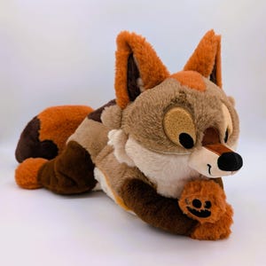 Puede incluir: Un peluche de zorro en tonos marrones, beige y naranja, tumbado sobre una superficie blanca. El zorro tiene orejas grandes, una nariz negra y una cara sonriente. Sostiene una peque&ntilde;a pata naranja con una cara de calabaza.
