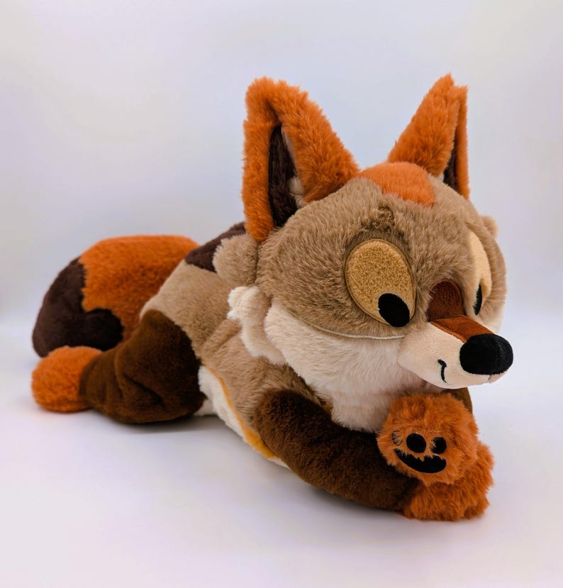 Puede incluir: Un peluche de zorro en tonos marrones, beige y naranja, tumbado sobre una superficie blanca. El zorro tiene orejas grandes, una nariz negra y una cara sonriente. Sostiene una peque&ntilde;a pata naranja con una cara de calabaza.