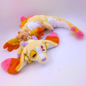 Könnte beinhalten: Ein skurriles Plüschtier in Gelb-, Orange- und Rosatönen. Das Spielzeug hat einen langen, segmentierten Körper und einen Kopf mit großen, ausdrucksstarken Augen. Ein kleiner Teddybär-Plüsch ist obenauf. Das Spielzeug ist weich und kuschelig, ideal für Kinder.