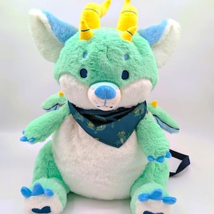 Może przedstawiać: Pluszowy smok w odcieniach zieleni, bieli i błękitu. Posiada żółte rogi, niebieskie oczy i niebieską bandanę we wzory. Smok ma miękką, futrzaną fakturę i jest zaprojektowany jako przytulanka.