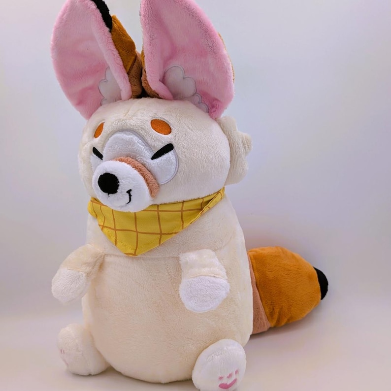 May include: A plush toy of a cartoon fox with large, pink-lined ears and a yellow bandana. The fox has a cream-colored body, orange eyes, and a brown and orange tail. The toy is sitting down with its paws visible.