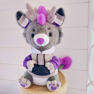 May include: A gray plush toy with purple and white accents. The toy has large ears with a white and purple stripe pattern, a purple mohawk, and a gray hoodie with a black pocket. The toy is sitting on a tan surface.