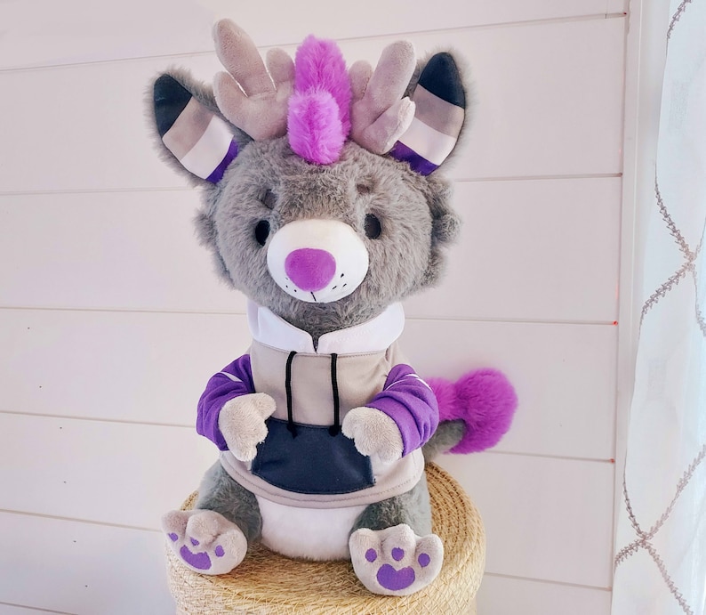 May include: A gray plush toy with purple and white accents. The toy has large ears with a white and purple stripe pattern, a purple mohawk, and a gray hoodie with a black pocket. The toy is sitting on a tan surface.