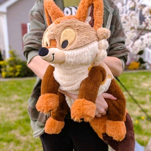 Puede incluir: Un peluche de zorro con pelaje marr&oacute;n, naranja y crema. El zorro tiene orejas grandes, una nariz negra y una expresi&oacute;n sonriente. El juguete se sostiene frente a un fondo borroso al aire libre.