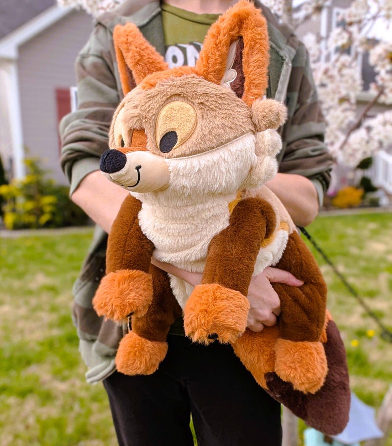 Puede incluir: Un peluche de zorro con pelaje marr&oacute;n, naranja y crema. El zorro tiene orejas grandes, una nariz negra y una expresi&oacute;n sonriente. El juguete se sostiene frente a un fondo borroso al aire libre.