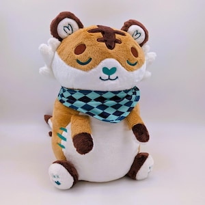 Peut inclure: Une peluche tigre avec une fourrure marron et blanche, une écharpe à carreaux bleus et noirs et un nez en forme de cœur. Le tigre a les yeux fermés et est assis. Le jouet a une queue et des pattes marron. La peluche mesure environ 25 cm de haut.
