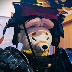 May include: A plush white dog wearing a black and red samurai helmet with gold accents. The dog has pink ears, orange eye markings, and a yellow collar. The armor is black and detailed.