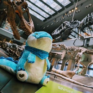 May include: A plush blue and cream-colored dinosaur toy with a blue plaid scarf, set against a museum backdrop of dinosaur skeletons. The toy has a heart-shaped detail on its foot.