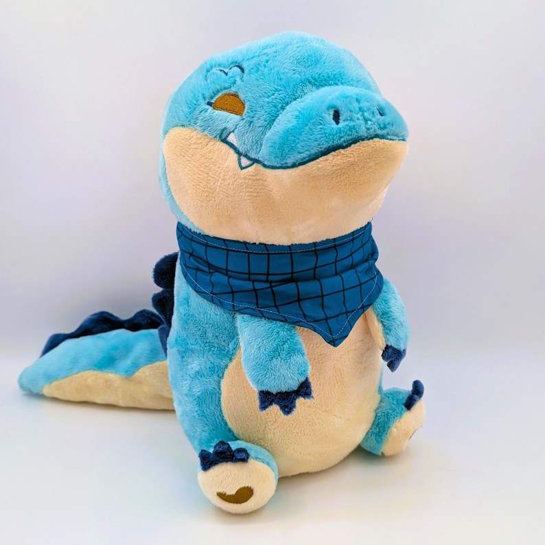 May include: A plush blue alligator toy with a light tan belly and a blue bandana. The alligator has a friendly expression and is sitting down with its tail extended. The toy is made of soft, plush material.