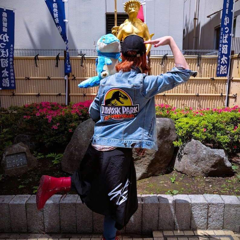 May include: A light blue denim jacket with a Jurassic Park logo patch on the back. The jacket is worn with a black skirt and red boots. A plush blue and white toy is on the person's shoulder.