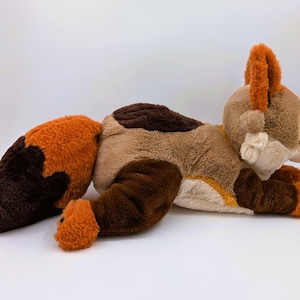 Puede incluir: Un peluche de zorro en tonos marr&oacute;n, beige y naranja, tumbado sobre una superficie blanca. El zorro tiene una larga cola, orejas puntiagudas y una cara detallada. El juguete es suave y tierno, perfecto para ni&ntilde;os.