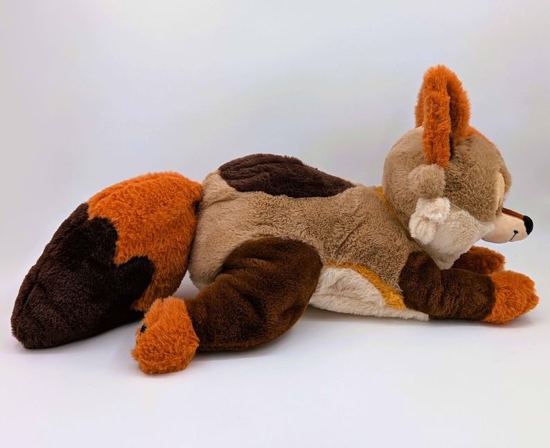 Puede incluir: Un peluche de zorro en tonos marr&oacute;n, beige y naranja, tumbado sobre una superficie blanca. El zorro tiene una larga cola, orejas puntiagudas y una cara detallada. El juguete es suave y tierno, perfecto para ni&ntilde;os.