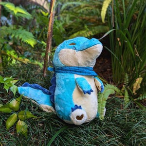 May include: A plush alligator toy in shades of blue and cream, with a blue scarf around its neck. The toy has a smiling expression and is set against a backdrop of green foliage.