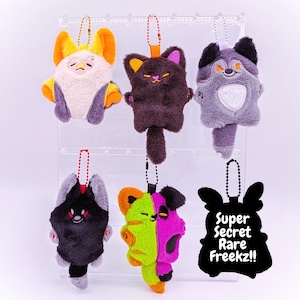 May include: A collection of plush keychains in various colors and designs, including a cat, a fox, and a split-faced creature. Each keychain has a metal chain and a small loop for attachment. The image also includes a black tag with the text "Super Secret Rare Freekz!!".