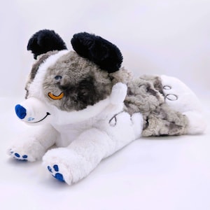 May include: A plush dog toy in shades of white, gray, and black. The toy has a smiling face with a blue nose and paw pads. The dog's ears are black, and the body is a mix of white and gray fur.