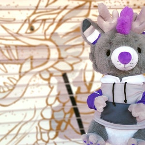 May include: A gray plush toy with purple accents, wearing a gray hoodie with purple accents. The toy has a purple nose, purple eyes, and a purple mohawk. The toy is sitting in front of a white and brown background.